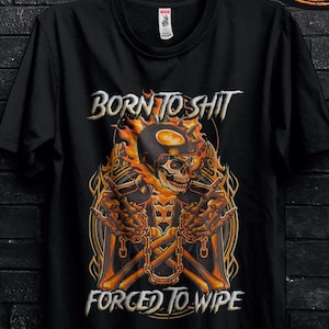 Può includere: T-shirt nero con un disegno di teschio con fiamme e la scritta "BORN TO SHIT FORCED TO WIPE". La maglietta è appesa a una gruccia nera. Il design è arancione e giallo.