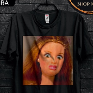 Può includere: T-shirt nero con il volto di una bambola con trucco esagerato. La maglietta presenta il logo "STITCH AURA" e testi promozionali: "50% OFF" e "BULK50". Il testo aggiuntivo recita "BUY 3 FREE" e "Code: SHIP3".