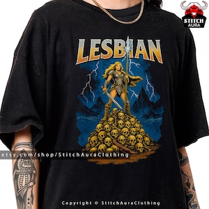 May include: Black t-shirt featuring the word "LESBIAN" in gold and orange. The graphic shows a female warrior with a spear and sword atop a pile of skulls, set against a backdrop of blue lightning.