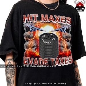 Puede incluir: Camiseta negra con el texto "HIT MAXES EVADE TAXES" en rojo y blanco. El diseño incluye aviones de combate, relámpagos y una pila de pesas de barra. Las pesas de barra están etiquetadas como "20,4 KG" y "45 LB".