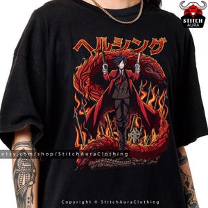 May include: Black t-shirt featuring a detailed anime-style illustration. The design includes a character in a red coat holding guns, set against a fiery dragon and Japanese text. Copyright StitchAuraClothing.
