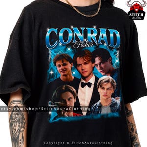 May include: Black t-shirt with a graphic design featuring several young men and the name "CONRAD Fisher" in a blue, stylized font. The design includes blue lightning bolts and stars, creating a vintage aesthetic. A casual wear item.