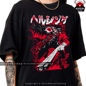 May include: Black t-shirt featuring a red and black graphic design with Japanese text. The design includes a stylized illustration of a character with a weapon and a wolf-like creature. The shirt has a crew neck and short sleeves.