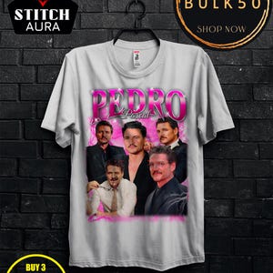 May include: A white t-shirt featuring a graphic of Pedro Pascal in various poses, with the name "PEDRO" in pink text. The shirt also has the text "STITCH AURA", "BUY 3 FREE", and "BULK50".