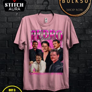 May include: A light pink t-shirt featuring a graphic of Pedro Pascal in various poses. The word "PEDRO" is in large, stylized letters above "Pascal". The shirt has a crew neck and short sleeves. The Stitch Aura logo is in the upper left corner.