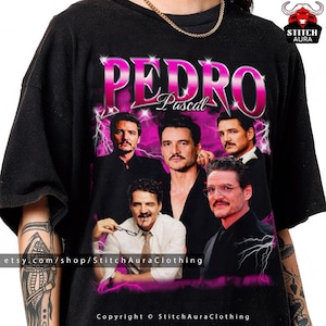 Puede incluir: Camiseta negra con un diseño gráfico rosa de Pedro Pascal y su nombre. El diseño incluye múltiples imágenes del actor con un fondo de rayos rosas. La URL de la tienda de Etsy y la información de derechos de autor están en la parte inferior.