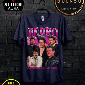 May include: Navy blue t-shirt featuring a graphic of Pedro Pascal with the name "Pedro" in large, pink letters. The shirt has a crew neck and is hanging on a black hanger. The image also includes the text "STITCH AURA", "BULK50", and "BUY 3 FREE".