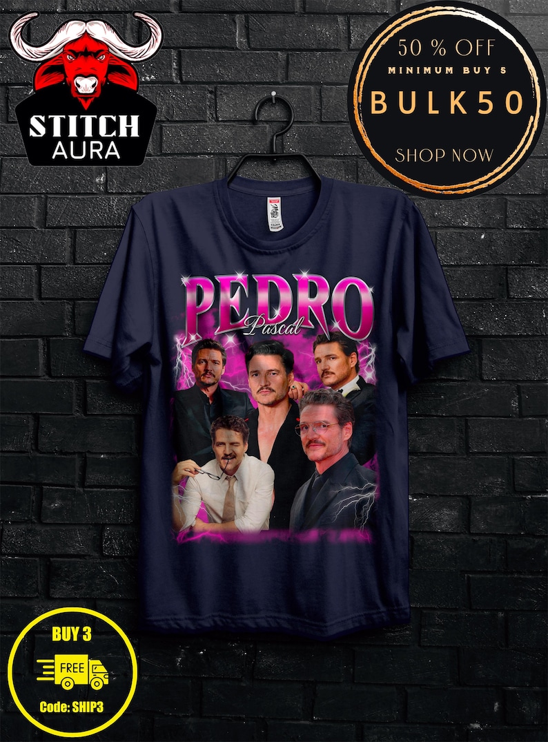 May include: Navy blue t-shirt featuring a graphic of Pedro Pascal with the name "Pedro" in large, pink letters. The shirt has a crew neck and is hanging on a black hanger. The image also includes the text "STITCH AURA", "BULK50", and "BUY 3 FREE".