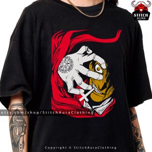 May include: Black t-shirt featuring a graphic design of a hand with a tattoo, red cape, and a stylized face with a cross. The design includes the text "Stitch Aura Clothing" and "Copyright © StitchAuraClothing".