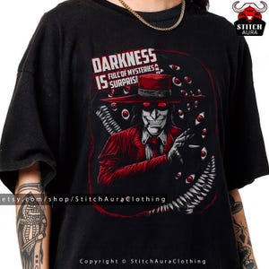 May include: Black t-shirt featuring a graphic of a character in a red suit and hat, with the text "DARKNESS IS SURPRISE" and "FULL OF MYSTERIES". The design includes red eyes and a sinister smile.