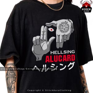 May include: Black t-shirt featuring a graphic design with two hands forming a frame around a red eye. The text "HELLSING ALUCARD" is in red and white, with Japanese characters below. Copyright StitchAuraClothing.