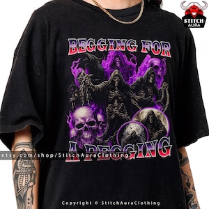 Puede incluir: Camiseta negra con un diseño gráfico que presenta calaveras, segadores y el texto "BEGGING FOR A PEGGIN". El diseño incorpora acentos morados y rojos, creando una estética oscura y gótica. La camiseta es una prenda casual.