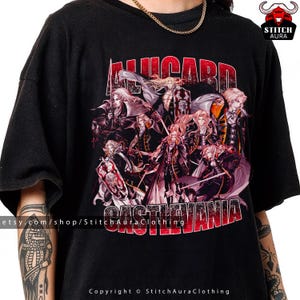 May include: Black t-shirt featuring a graphic design with the text "ALLUCARD CASTLEVANIA" in red and white. The design includes multiple characters in dark clothing with long hair. The shirt has a crew neck and short sleeves.