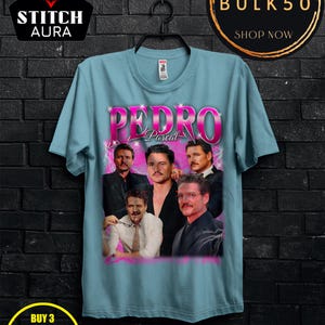 May include: Light blue t-shirt featuring a graphic of Pedro Pascal with the name "Pedro Pascal" in pink. The shirt has a crew neck and short sleeves. The image includes the "STITCH AURA" logo and promotional text.