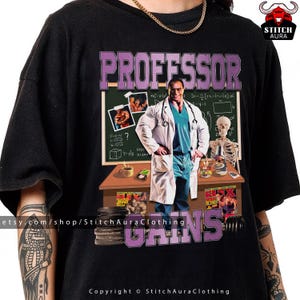 Pode incluir: Camiseta preta com um gráfico de um fisiculturista em jaleco de laboratório e o texto "PROFESSOR GAINS". O design inclui um quadro-negro, um esqueleto e pesos. A camiseta tem gola redonda.