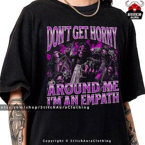 Könnte beinhalten: Schwarzes T-Shirt mit einem lila Grafikdesign, das Skelettfiguren mit Waffen zeigt, mit dem Text "DON'T GET HORNY AROUND ME I'M AN EMPATH". Die Etsy-Shop-URL und Copyright-Informationen sind ebenfalls sichtbar.