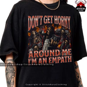 Può includere: T-shirt nera con grafica di figure scheletriche con pistole, fiamme e la scritta "DON'T GET HORNY AROUND ME I'M AN EMPATH." La maglietta include il nome del negozio Etsy e le informazioni sul copyright.