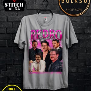 May include: Gray t-shirt featuring a collage of images of Pedro Pascal with the name "PEDRO" in pink, and the word "Pascal" below. The shirt has a crew neck and short sleeves. The shirt is hanging on a black hanger.