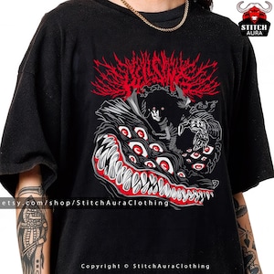 May include: Black t-shirt featuring a detailed graphic design. The design includes a red, stylized, spiky element above a creature with multiple red eyes and sharp teeth. The text "STITCH AURA" is visible on the upper right.