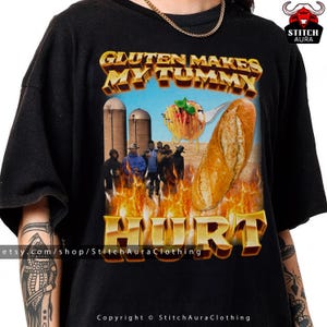 May include: Black t-shirt with a graphic design featuring the text "Gluten Makes My Tummy Hurt" in gold lettering. The design includes a baguette, pasta, and a group of people in front of a farm scene.