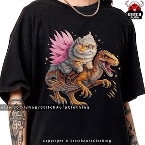 Può includere: T-shirt nero con un'illustrazione stravagante di un gatto con occhiali e una corona, con ali di piume rosa, in sella a un dinosauro. Il design include stelle e il testo "Stitch Aura Clothing".