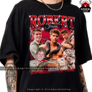 May include: Black t-shirt with a graphic of Robert Irwin, a snake, crocodile, and koala. The name "ROBERT Irwin" is in red, stylized text. The shirt includes the text "Copyright © StitchAuraClothing".