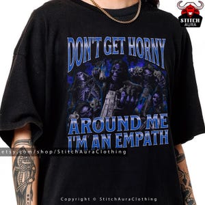Könnte beinhalten: Schwarzes T-Shirt mit dem blauen Text "DON'T GET HORNY AROUND ME I'M AN EMPATH". Die Grafik zeigt Skelettfiguren mit Waffen. Die Etsy-Shop-URL und Copyright-Informationen befinden sich unten.