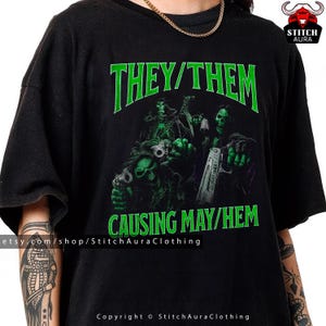 May include: Black t-shirt with a green graphic of skulls holding guns, and the text "THEY/THEM CAUSING MAY/HEM". The Etsy shop URL and copyright information are visible at the bottom of the shirt.