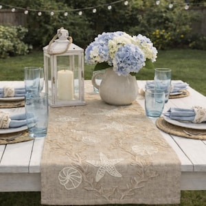 Op de afbeelding: Een witte houten tafel gedekt voor een maaltijd in de buitenlucht met een maritiem thema. Een beige tafelloper met geborduurde zeesterren en schelpen siert de tafel. Blauwe servetten, glazen en een vaas met hortensia's maken het geheel af. Een witte lantaarn met een kaars is ook aanwezig.