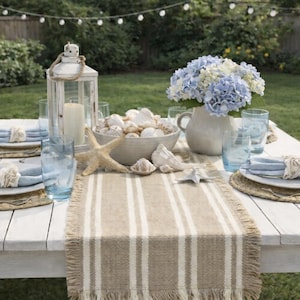 May include: A white wooden table set for an outdoor meal. The table is decorated with blue napkins, plates, and seashell accents. A vase of blue and white hydrangeas and a white lantern complete the coastal theme.