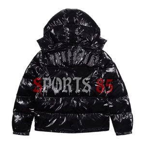 Pode incluir: Casaco puffer preto com capuz. O casaco tem a palavra "TRAPSTAR" nas costas e "SPORTS $5" em fonte g&oacute;tica. O casaco &eacute; brilhante e tem um design acolchoado.