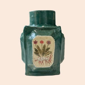 May include: A teal ceramic vase with a rectangular body and a wide neck. The vase has a central panel with a botanical illustration of flowers and roots. The panel is framed with small, white dots. The vase is handmade.