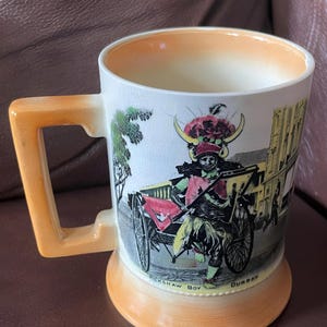 May include: A vintage ceramic mug featuring a detailed illustration of a rickshaw boy in Durban. The mug has a cream-coloured body with an orange handle and base. The illustration includes a rickshaw, figures, and text. The mug's rim and interior are also orange.