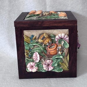 Op de afbeelding: Een donkerbruine houten kist met een gedetailleerde reliëfscène van een tuin. Het reliëf toont bloemen, insecten en kleine dieren. De bovenkant van de kist heeft een reliëf van kleine dieren. Het is een decoratief item.