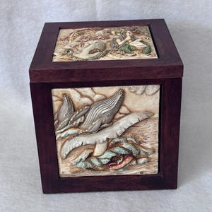 Op de afbeelding: Een vierkante houten doos met een donkerroodbruin frame en gedetailleerde reliëfsnijwerken. De snijwerken tonen walvissen, zeemeerminnen en andere zeedieren in crème. De doos heeft een deksel.