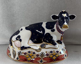 Puede incluir: Figura de vaca de cerámica en posición reclinada. La vaca es blanca con manchas negras y una nariz dorada. La base presenta un diseño floral rojo, dorado y azul. La vaca tiene un collar rojo.