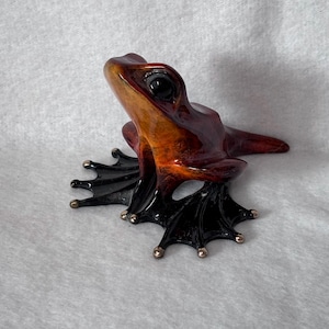 May include: A detailed sculpture of a frog with a glossy finish. The frog's body is a blend of orange and brown hues, with black feet and a dark eye. The feet have small, silver-colored accents.