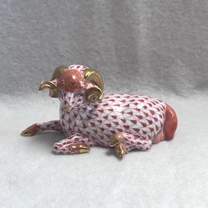 May include: A ceramic ram figurine in a resting pose. The ram features a red and white pattern, with gold-colored horns and accents on the hooves. The figurine is a decorative item.