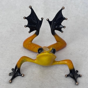 May include: A small frog figurine with a bright yellow head and body that transitions into orange and black limbs. The frog is depicted in a leaping pose with its back legs extended upward, featuring black webbed feet with small metallic tips.
