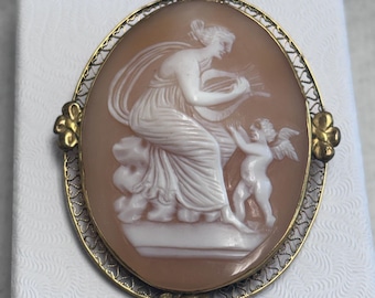 Broche/Colgante antiguo de filigrana chapada en oro con camafeo de concha que representa a una musa clásica tocando una lira, acompañada por un juguetón Cupido.
