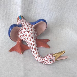 May include: A ceramic duck figurine with a white body, red and white patterned neck, and red feet. The duck has blue wings, a gold beak, and is lying on its side. The figurine is a decorative item.