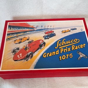 May include: A red box with a vintage illustration of race cars on a track. The illustration features a red race car with the number 17 in the foreground, and the text "Schuco Grand Prix Racer 1075". The box has a red border.