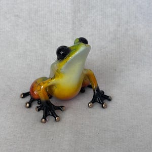 May include: A small frog figurine with a smooth, glossy finish. The body features a gradient of bright yellow and orange, transitioning to deep black on the limbs and feet. It has large, prominent black eyes and small, rounded toe pads.