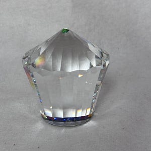 Puede incluir: Objeto de cristal transparente y facetado, con forma de diamante. Tiene una base plana y una parte superior puntiaguda con un pequeño acento verde. Las facetas crean un juego de luz y color.