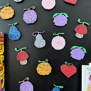 Op de afbeelding: Een verzameling kleurrijke, gestileerde fruitvormige magneten. De magneten hebben verschillende ontwerpen, waaronder hart-, peer- en bloemvormen, in tinten rood, paars, geel, blauw en roze. Elke magneet heeft een steel en is gerangschikt op een zwart oppervlak.