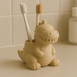 Op de afbeelding: Een beige dinosaurusvormige tandenborstelhouder met twee tandenborstels. Eén tandenborstel is wit en de andere is bruin. De houder heeft een lachend gezicht en is gemaakt van een glad, mat materiaal. De houder is ontworpen om tandenborstels rechtop te houden.