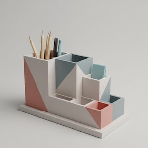 Pode incluir: Um organizador de mesa geométrico em branco, cinza e coral. O organizador tem vários compartimentos para canetas, lápis e objetos pequenos. Lápis e canetas são visíveis. O design é moderno e minimalista.
