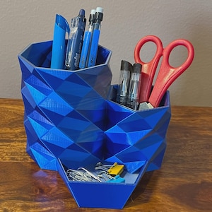 May include: A geometric blue desk tidy with multiple compartments. It holds pens, pencils, scissors, paper clips, and other stationery. The organiser is made of a textured, faceted material and sits on a wooden surface.