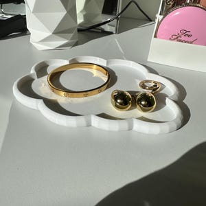 May include: A white cloud-shaped tray holds gold jewelry: a bracelet, a pair of earrings, and two rings. The tray sits on a white surface, with a white geometric container and makeup products in the background.