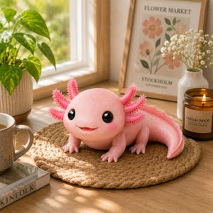 Puede incluir: Una figura de ajolote de color rosa claro con grandes ojos negros, una boca sonriente y prominentes branquias externas rosas. La criatura tiene una textura suave y moteada y está colocada sobre un mantel individual redondo de color beige, mostrando sus cuatro extremidades.